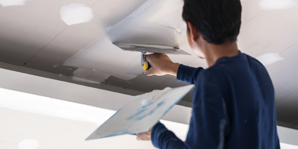 Construction workers installing drywall panels on a ceiling with metal framing.