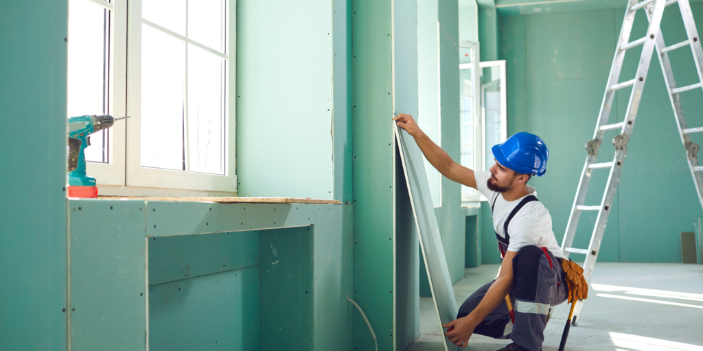 Construction worker installing green moisture-resistant drywall panels in a commercial building.
